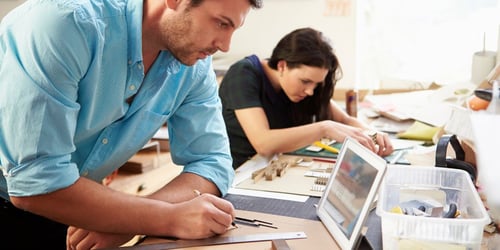 Man writing a note while looking at an iPad; women in background working