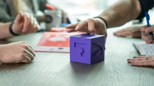 Purple dice on table with a hand touching it.